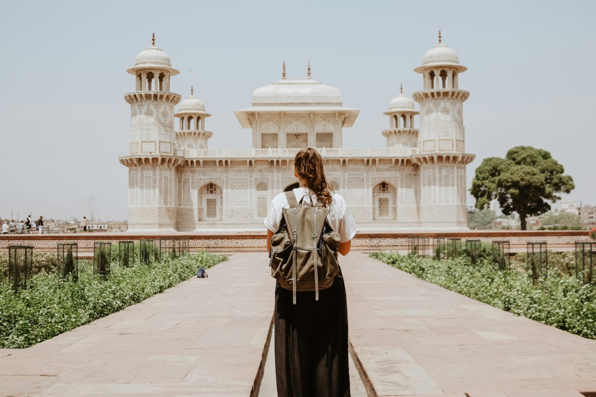 A traveler with a backpack stands facing an ornate white tomb, surrounded by gardens under a clear sky.